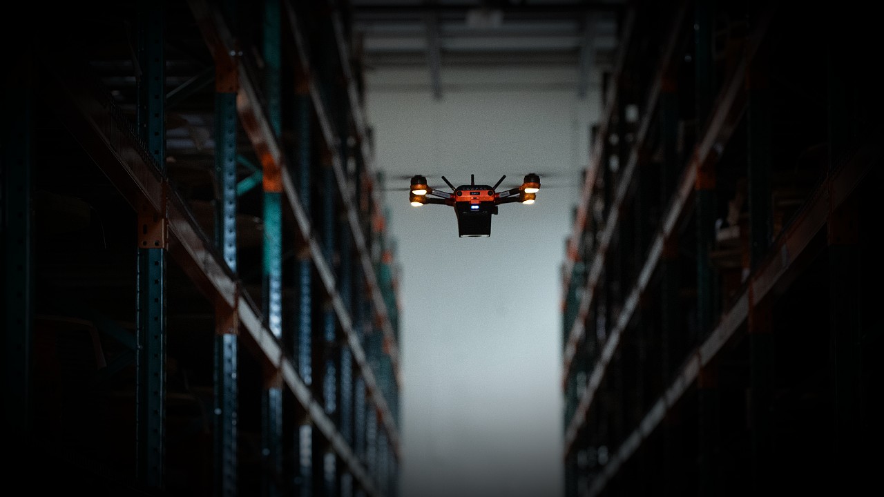 A Corvus One warehouse drone flies through a dark aisle as it scans inventory.